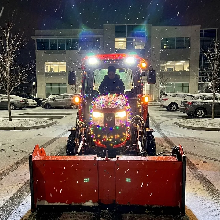 Tracteur de déneigement illuminé qui déblaie le stationnement d'un édifice de bureaux la nuit sous la neige, voitures stationnées de chaque côté