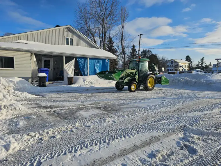 Tracteur vert avec lame de déneigement au bout d'une entrée dégagée, entre de hauts bancs de neige, par temps clair